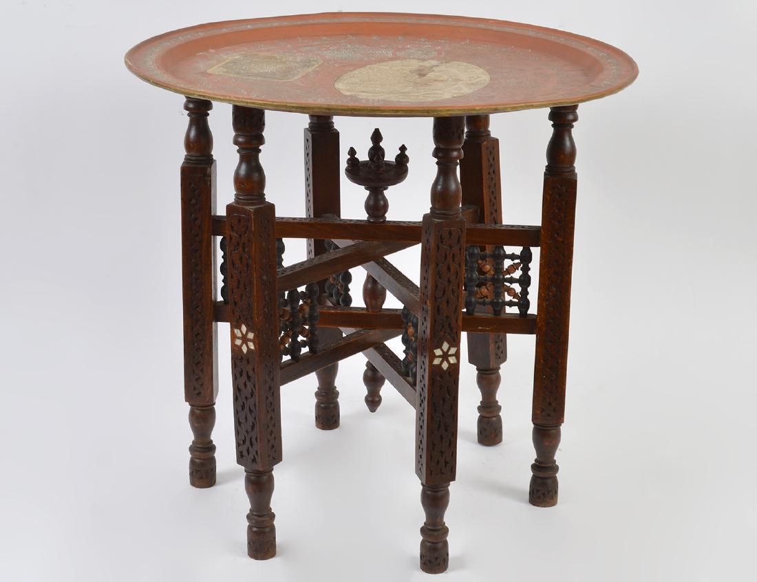 MIDDLE EASTERN BRASS TRAY ON STAND: The circular tray incised with stylized Islamic script and desert motifs, on a bone inlaid wood folding stand. Height 20”. Diameter 21”. (Cond: tray with surface blemishes, stand nice)