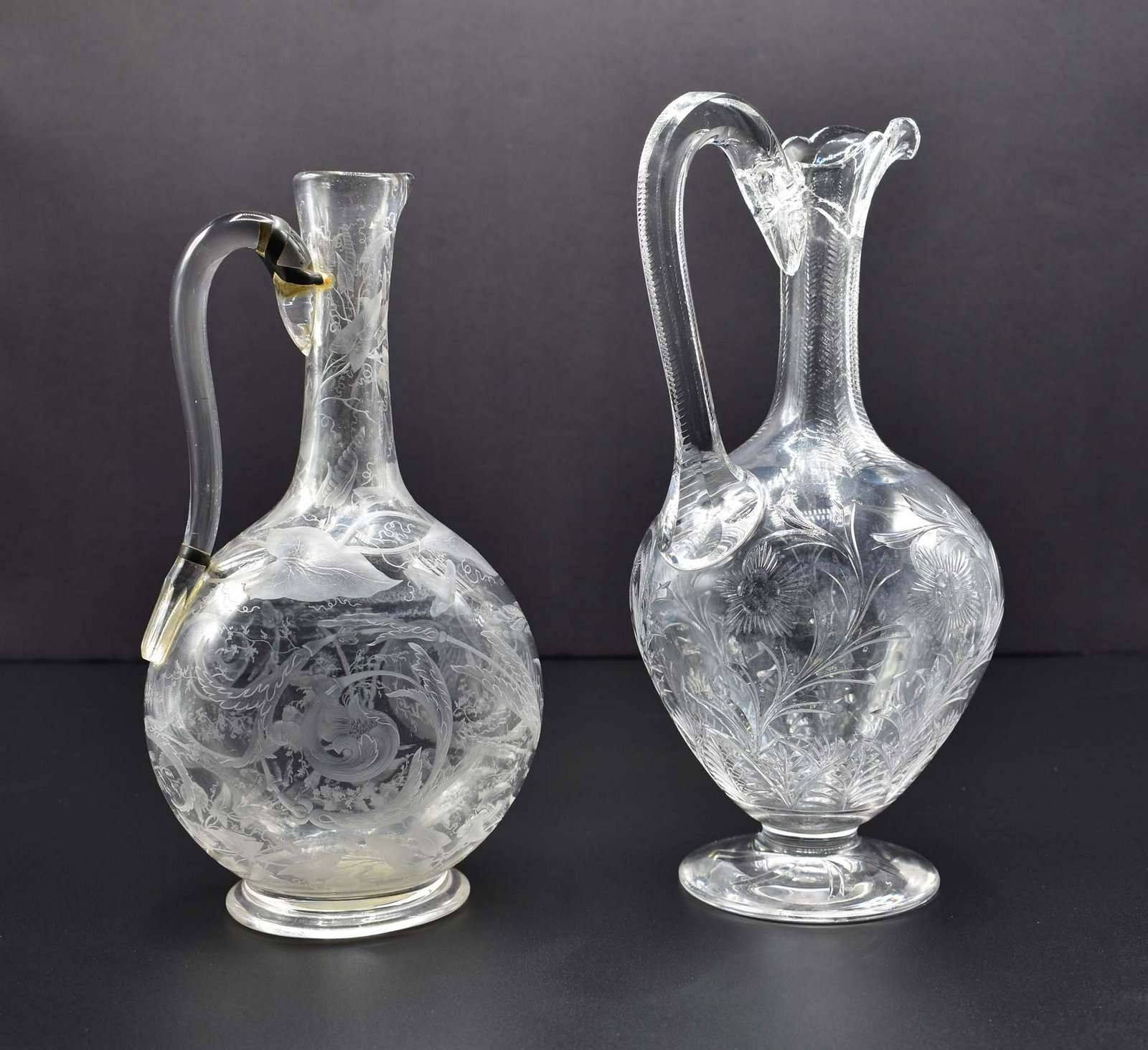 TWO ENGLISH WHEEL-ENGRAVED COLORLESS GLASS EWERS: Both Late 19th Century. The first Stourbridge, with a loop handle and engraved with flowers, the second, probably Lobmyer, engraved with a classical satyr and scrolling foliage. Height 10.5 in., 9.75 
