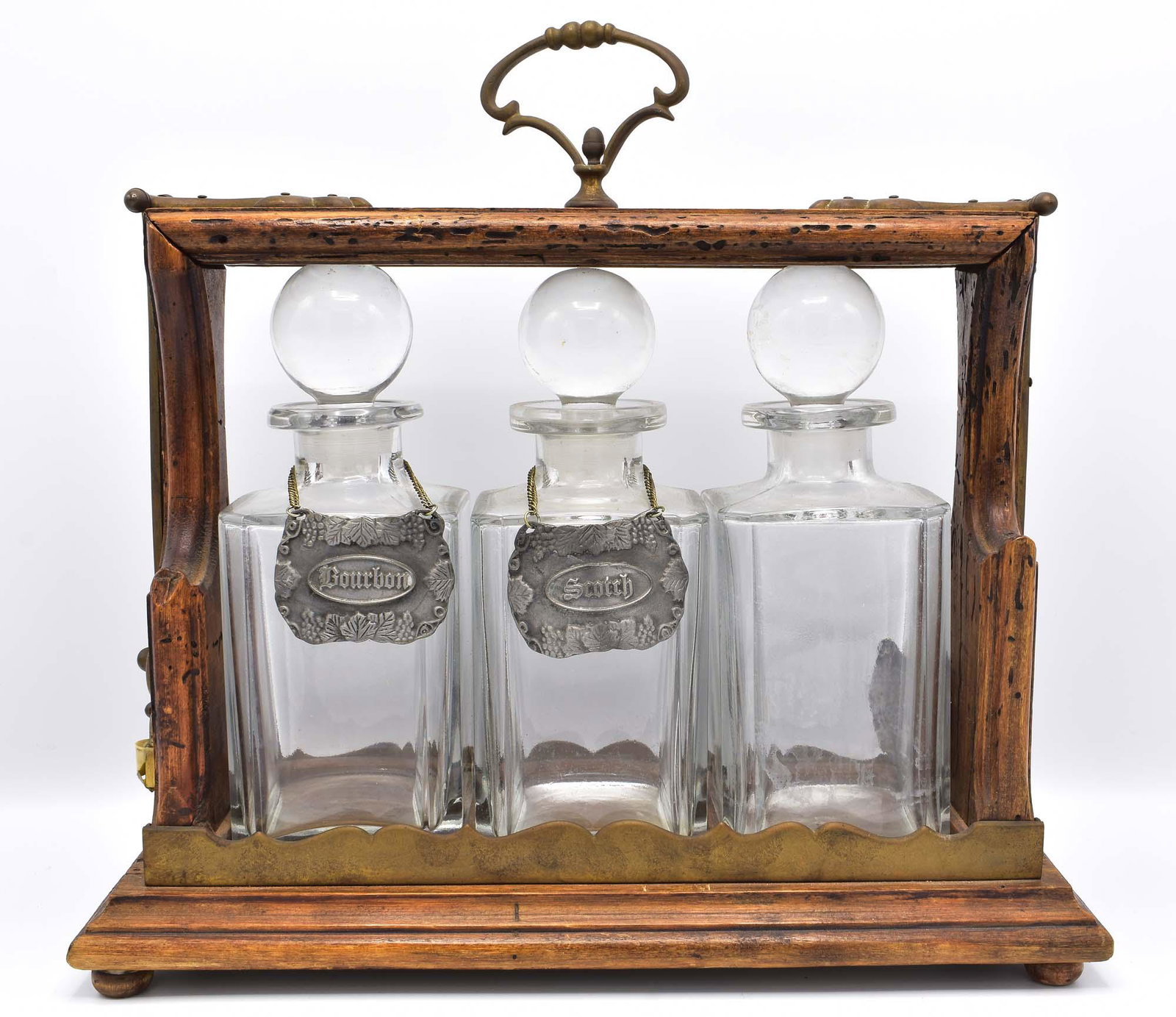 BRASS MOUNTED OAK TANTALUS & THREE GLASS DECANTERS: Early 20th Century. Probably American. The case with a locking hinged side, the three decanters with spherical stoppers and faceted corners. Height 14 in. Width 14.5 in. (Cond: good)