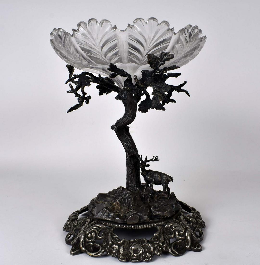 SILVERED METAL AND CUT GLASS CENTERPIECE: Probably English, Circa 1900. The rockwork with illegible hallmarks, also etched with an eagle above an inscribed banner. Cast as a leafy tree on a rocky mound, a deer at the base, supporting a colorl