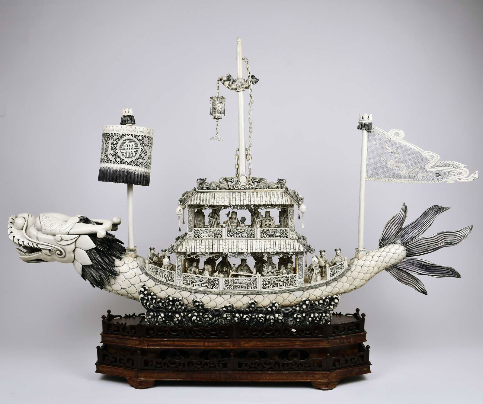 MASSIVE CHINESE CARVED DRAGON BOAT ON STAND: The scale carved dragon-headed boat, with a pierced and tiered deck, with fret galleries, crowded with passengers engaged in leisurely pursuits, the deck fitted with a pierced cylindrical lantern, and