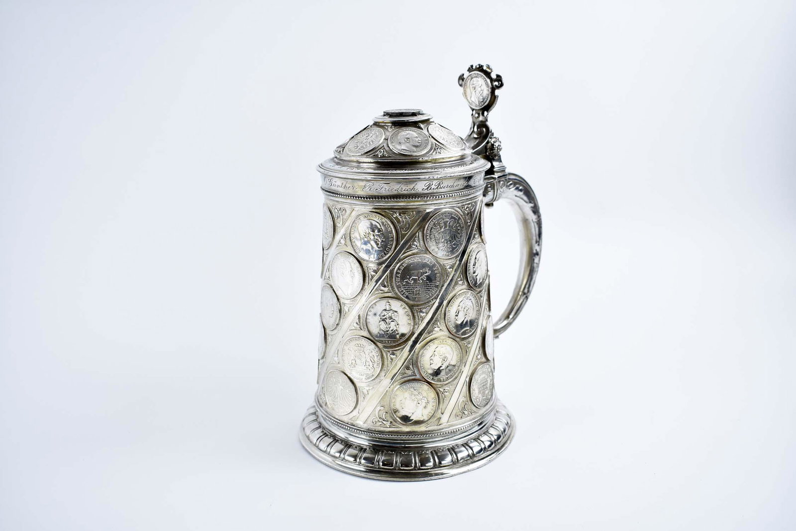 CONTINENTAL COIN-INSET SILVER PLATE TANKARD: Late 19th Century. The cylindrical vessel with hinged lid and loop handle, the lid with a thumb flange, the body inset with silver and plated coins and medallions, many with portraits and names, some 