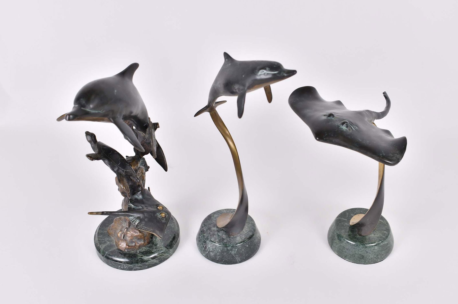 THREE PATINATED BRONZE MARINE LIFE SCULPTURES: Modern. No visible markings. Depicting a stingray, a dolphin and a group with a dolphin swing amongst a turtle and ray, each atop either a twisting bronze support or coral group, on a marble base. Hei