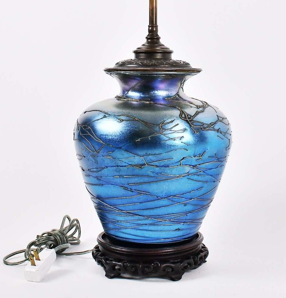 BLUE IRIDESCENT GLASS VASE MOUNTED AS A LAMP: Possibly American. The tapered spherical vessel with raised linear motifs, with a stamped metal cover cap and carved wood Chinese base, drilled. Height 10 in. Diameter at shoulder 9 in. Height overall
