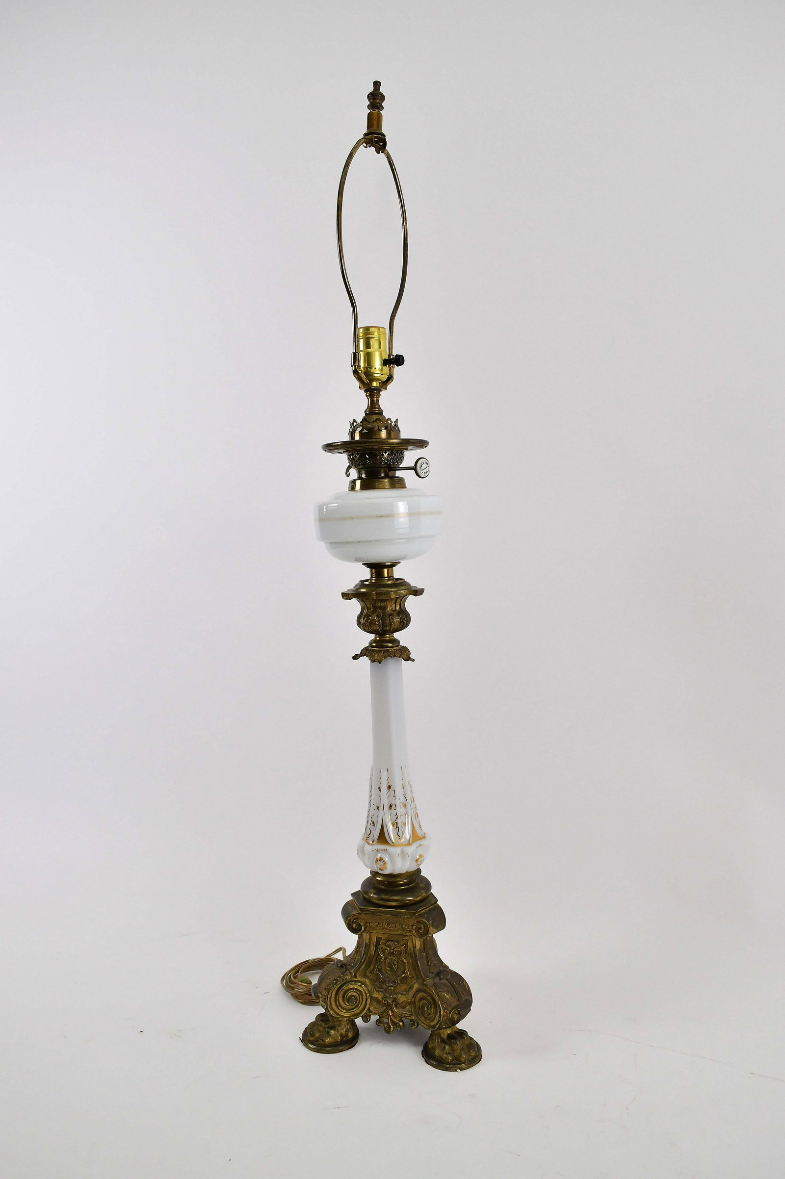 VICTORIAN OPALINE AND BRASS OIL LAMP: Second Half 19th Century. The repousse decorated tripod base supporting a parcel gilt white glass standard, beneath a glass reservoir, electrified. Height 27 in. (Cond: adapted)