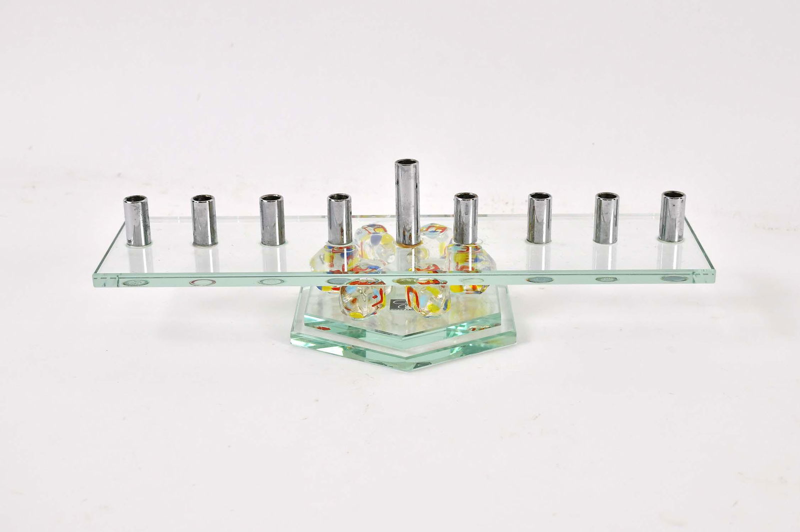 CONTEMPORARY GLASS MENORAH: Chromed candle holders on a glass platform supported on six colorful dreidels, set upon a hexagonal base. Width 12.25 in.