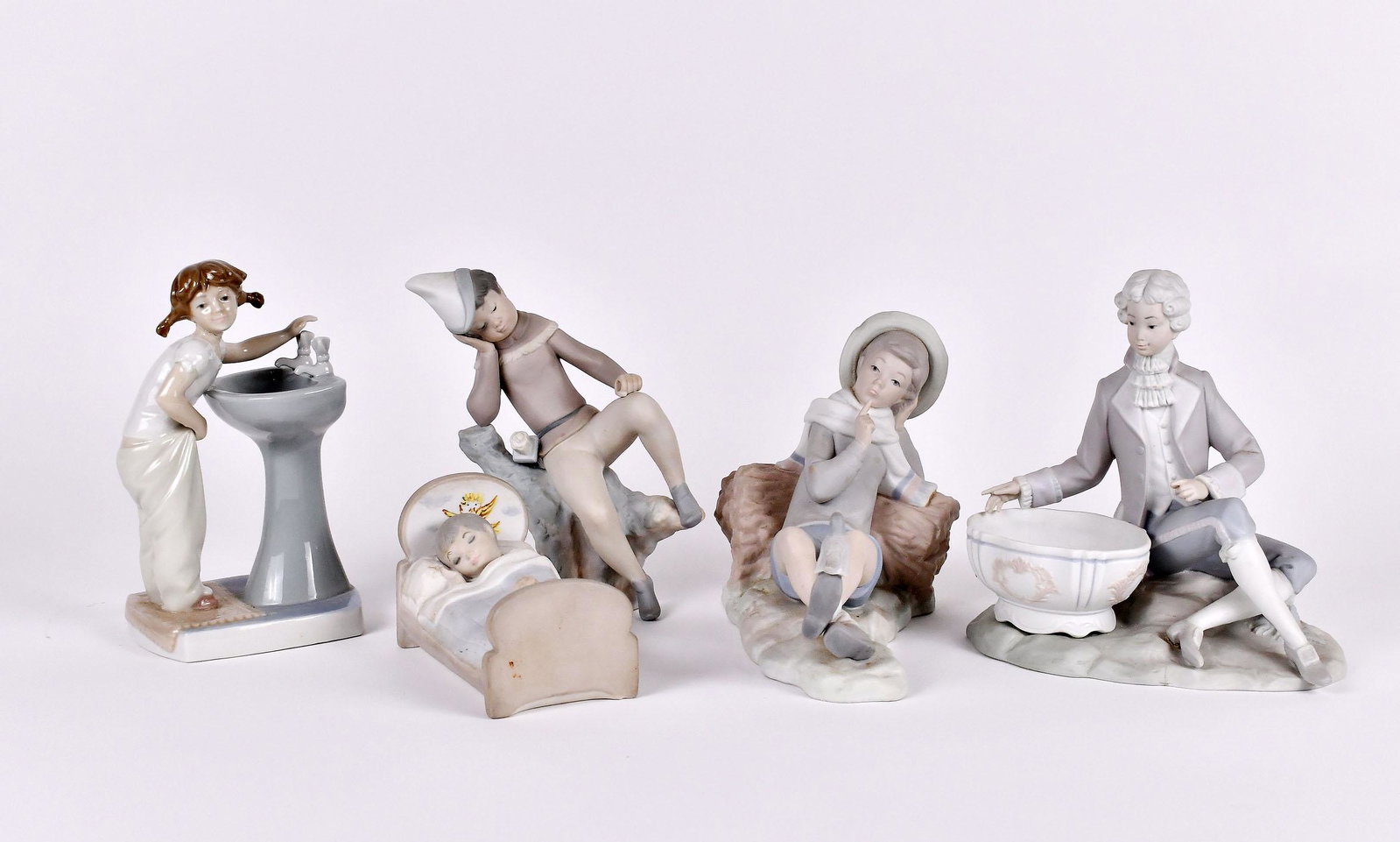 FIVE LLADRO PORCELAIN FIGURES: Each signed, numbered on the underside. Comprising: a gentleman in 18th Century attire seated before a basin, a girl washing at a sink, a boy asleep in his bed, a boy reclining on a bundle of sticks a