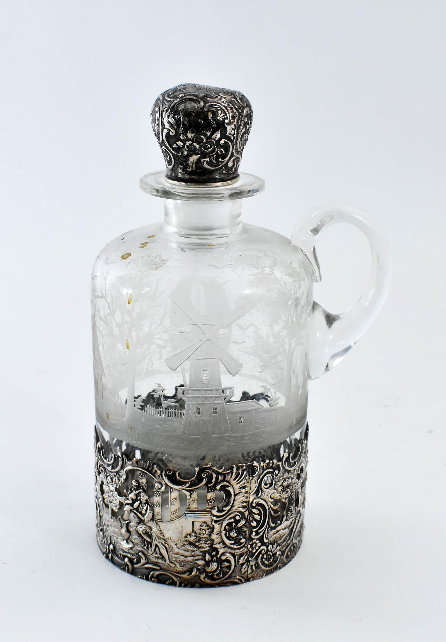 DUTCH SILVER MOUNTED INTAGLIO GLASS DECANTER: Marked, maker SVK. Of cylinder shape, the glass decorated with a village and windmills, the silver chased figures in a rococo garden, with a loop handle and silver covered stopper. Height 8.5 in. (Con