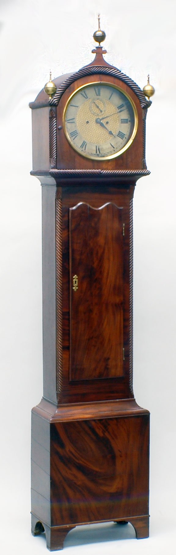 GEORGE III MAHOGANY TALL CASE CLOCK: GEORGE III MAHOGANY TALL CASE CLOCK English. Circa 1805. By John Warren Canterbury. The circular dial with Roman numerals signed Warren Canterbury housed within the hood with finials and glazed door,