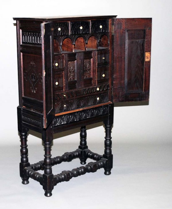 CHARLES II STYLE CARVED OAK SIDE CABINET ON STAND: English. 18th/19th Century. The rectangular top above a pair of cupboard doors opening to the interior fitted with drawers, pigeon holes and a pair of paneled doors over two long drawers, the base wit