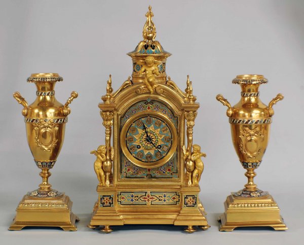 THREE PIECE GILT BRONZE AND CHAMPLEVE ENAMEL CLOCK: French. 19th Century. Comprising: clock and a pair of urns. The architectural case centered by the dial with Roman numerals surmounted by a seated putto and a cupola, each corner with a post and two s