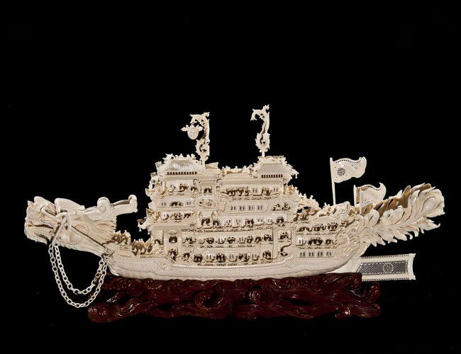 HIGHLY DETAILED CARVED IVORY ROYAL DRAGON BOAT: Chinese. The seven level vessel with dragon mast head and two detachable anchors, fitted with a multitude of passengers and various lanterns and banners. Length 24 ½". On a carved wood stand. Overall
