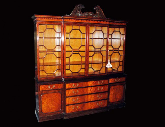 GEORGE III STYLE MAHOGANY BREAKFRONT: American. By Schmieg & Kolzian Inc., N.Y. In two parts. The upper with pierced swan neck cresting above a dentil cornice and four glazed doors, the lower with a silver drawer, two slide tablets, two s
