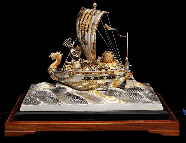 SILVER AND SILVER GILT MODEL OF A TREASURE SHIP: Japanese. The vessel with bird's mast under full sail carrying its treasure trove over naturalistic waves. Length 11". Height 7 ¾". Depth 6 ¾". In a glass case. With dedication plaque "with the comp