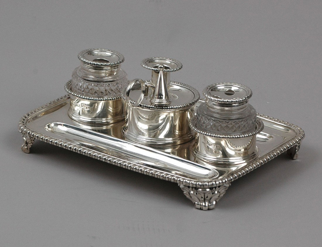 GEORGE III STERLING SILVER STANDISH: English. Birmingham. 1818. Maker: Matthew Boulton. The rectangular stand with gadroon border and two pen wells and fitted two glass inkwells and centered by a chamber stick with snuffer. Length 9" (23