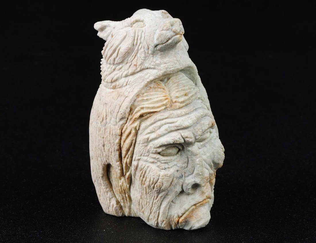CARVED STONE HEAD OF A SHAMAN: Inuit. Indistinctly signed and dated '01. Of an elderly Shaman covered by a wolfskin cloak. With engraved paw print on reverse. Height 4 1/8". (100/150)