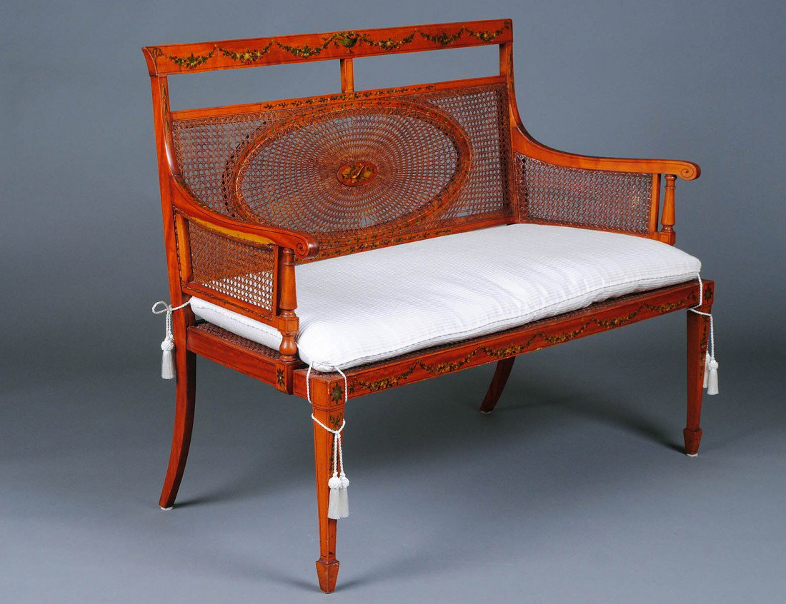 EDWARDIAN PAINTED SATINWOOD LOVE SEAT: English. Circa 1910. With cane backrest and seat flanked by cane arms, raised on square tapered legs ending in spade feet. Length 50 3/4". (1500/2000)