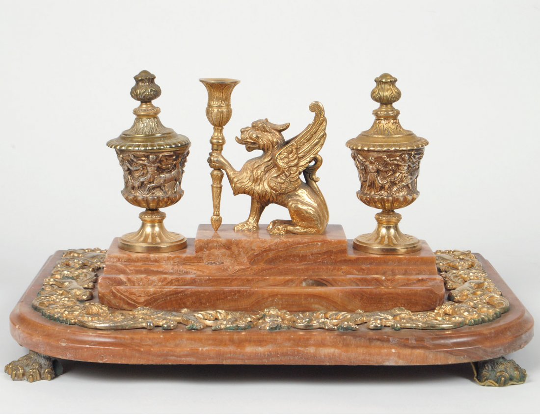 GILT BRONZE MOUNTED ONYX DESK STAND: The D-shaped base supporting pen wells and urn-form inkwells centered by a winged lion candle holder, the whole raised on paw feet. Length 17 1/2". (600/800)
