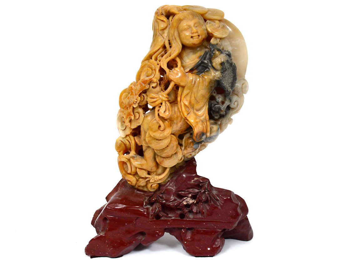 CARVED SOAPSTONE FIGURE OF QUANYIN: Chinese.The standing deity holding a string with coins and a fish flanked by fungi and a bat, raised on a soapstone base. Height 10". (200/300)