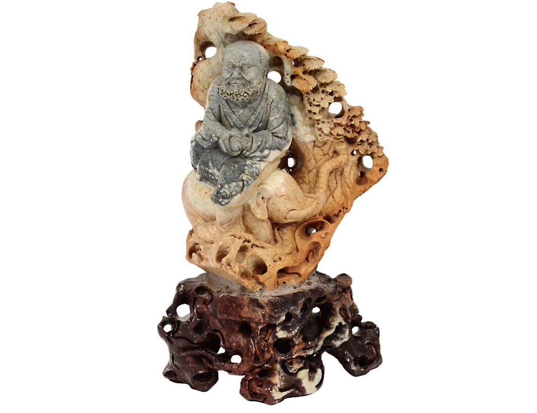 CARVED SOAPSTONE FIGURE OF A LOHAN: Chinese. Seated on an elephant under a pine tree, raised on a soapstone base. Height 11". (200/300)