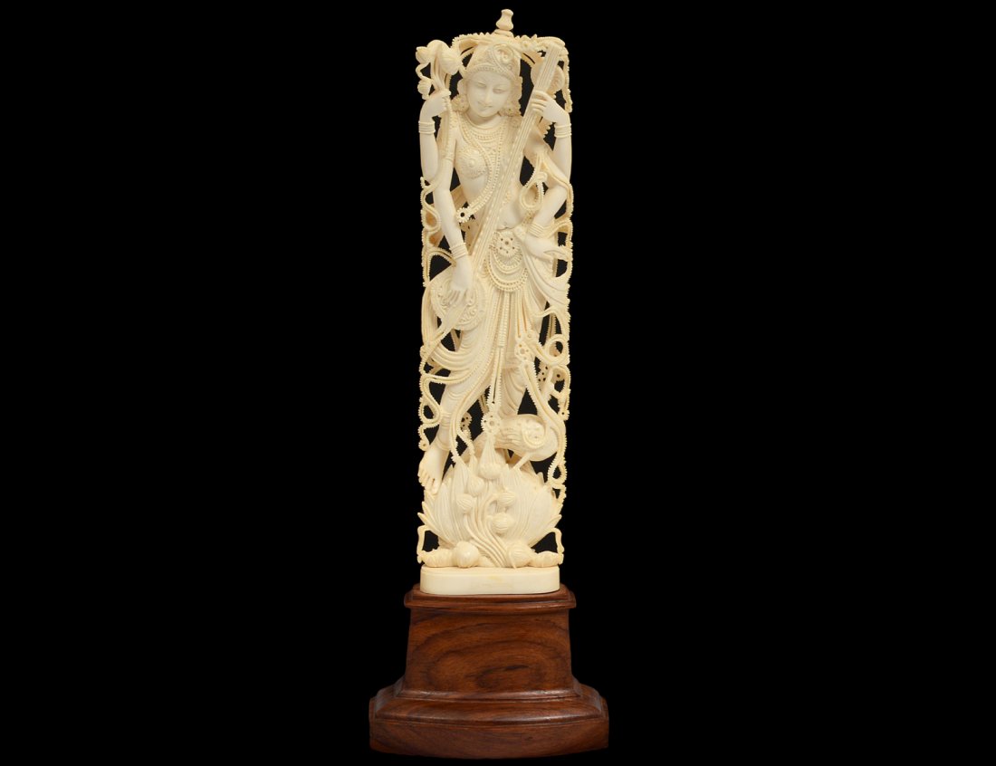 ELABORATELY CARVED IVORY FIGURE OF A FEMALE MUSICIAN: Indian. Standing on a blossom with a bird holding a string instrument. Height 12". On a wood base. (800/1200)