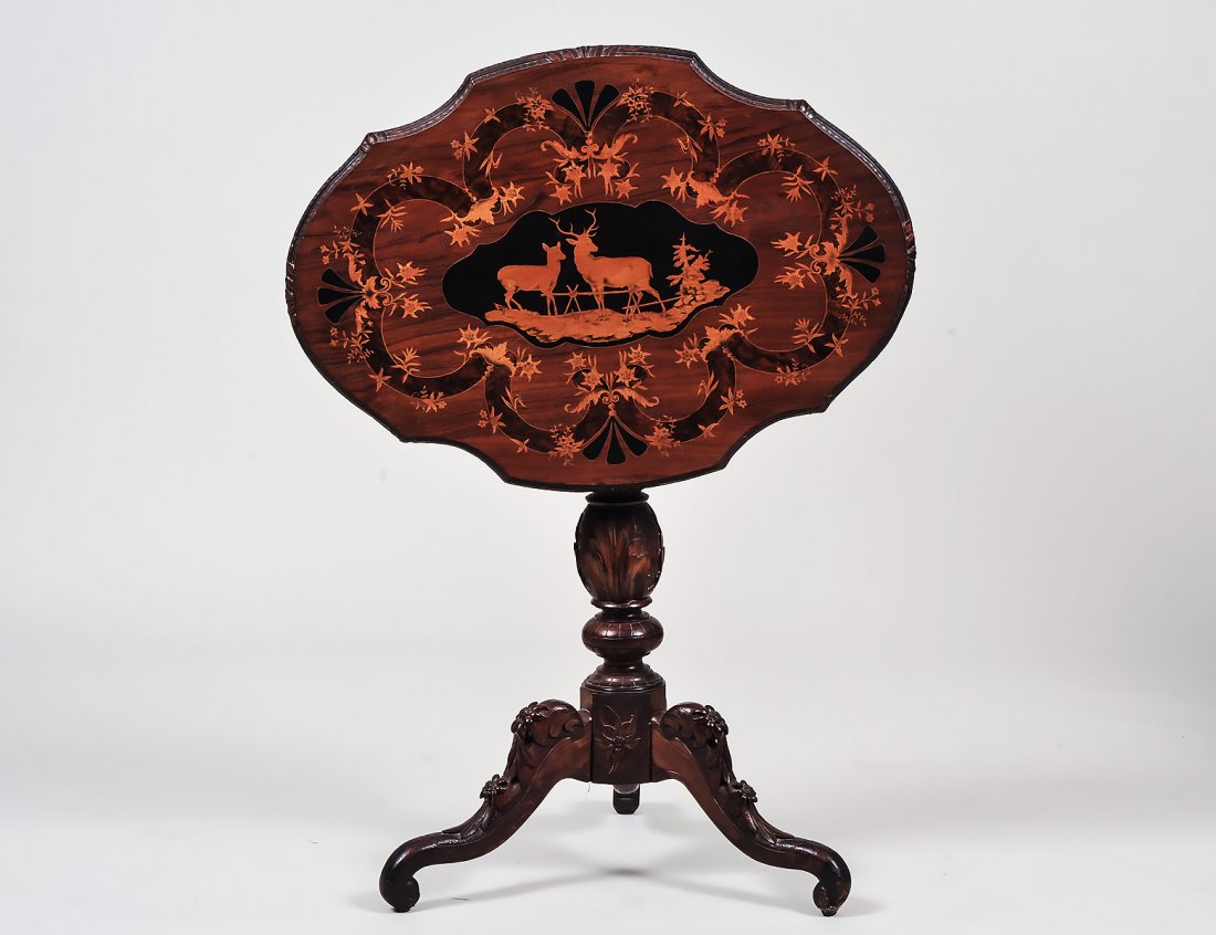BLACK FOREST INLAID WALNUT TILT-TOP TRIPOD TABLE: Late 19th Century. The shaped oval foliate carved top also inlaid with two deer, foliage and scrolls, on a turned foliate carved standard, on similarly carved shaped legs ending in paw feet. Height 28