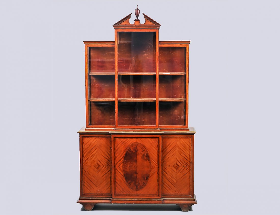 GEORGE III INLAID SATINWOOD BOOKCASE CABINET: In the manner of Thomas Sheraton. In three parts: the triangular pediment above three glazed doors. The conforming projecting lower part with a brass inset edge, fitted with a single oval inlaid door,