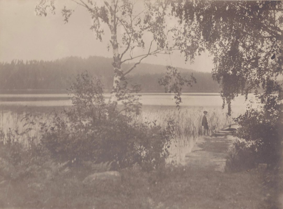 A Young Boy Amidst Scandinavian Landscape, 1912.: Sepia-tone photograph showing a young boy and rowboat surrounded by a Scandinavian landscape. Stamped "Nanna Ostman, Nykoping, 1912" on recto. Inscribed and dated in period handwriting on verso. Mediu