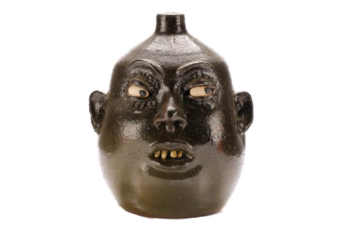 Lanier Meaders Face Jug, Four Teeth, Signed: Lanier Meaders (American, White County, Georgia, 1917-1998). A face jug in an uneven olive glaze, with incised decoration, painted black eyes, an open mouth with four teeth, and an applied strap handl