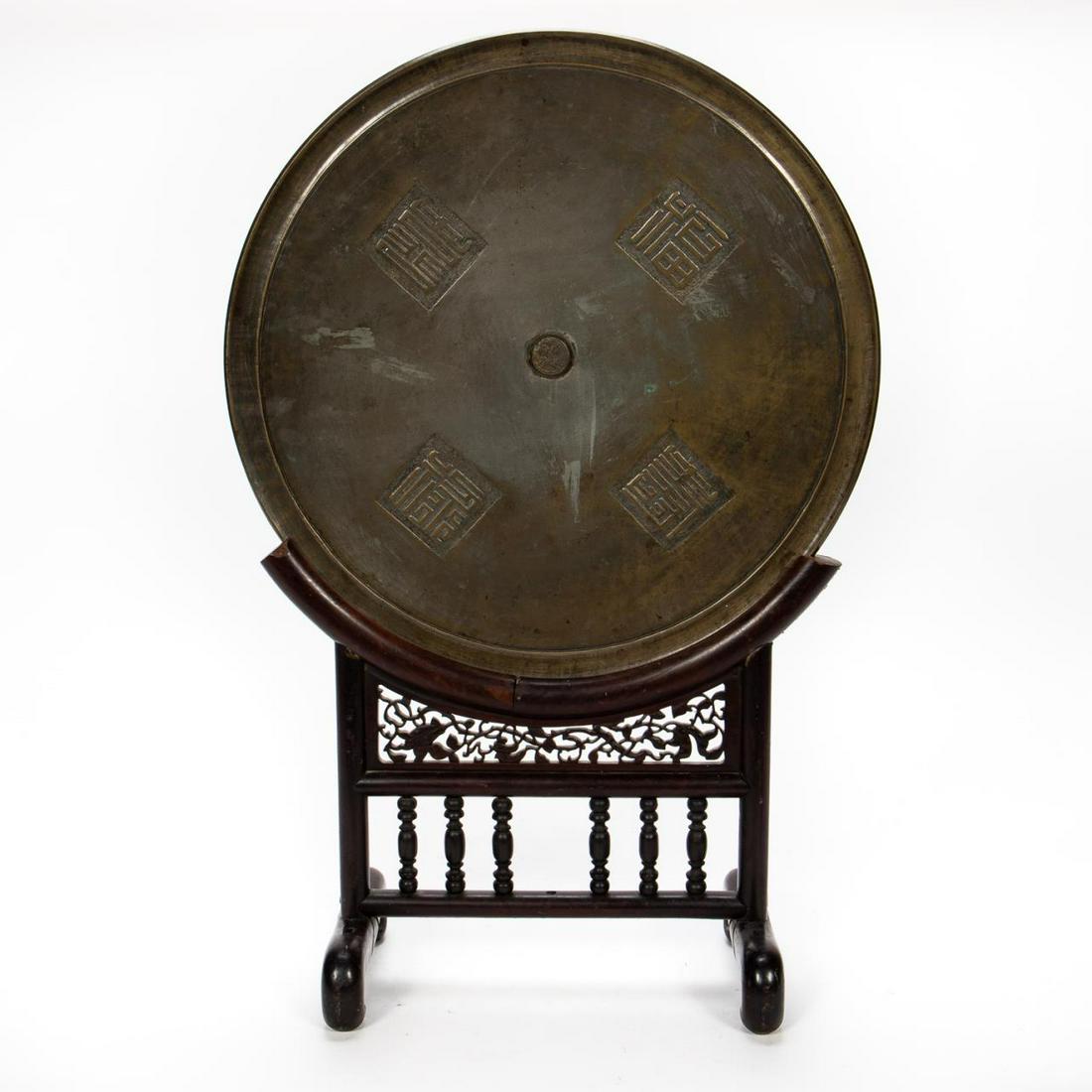 Chinese Bronze Mirror on Rosewood Stand: Chinese, circa 1900. Circular cast bronze mirror decorated with four characters, on a rosewood stand with fretwork and turned spindles. Signed to center. Approx. on stand h. 30.5", w. 20.25", d. 8.5";