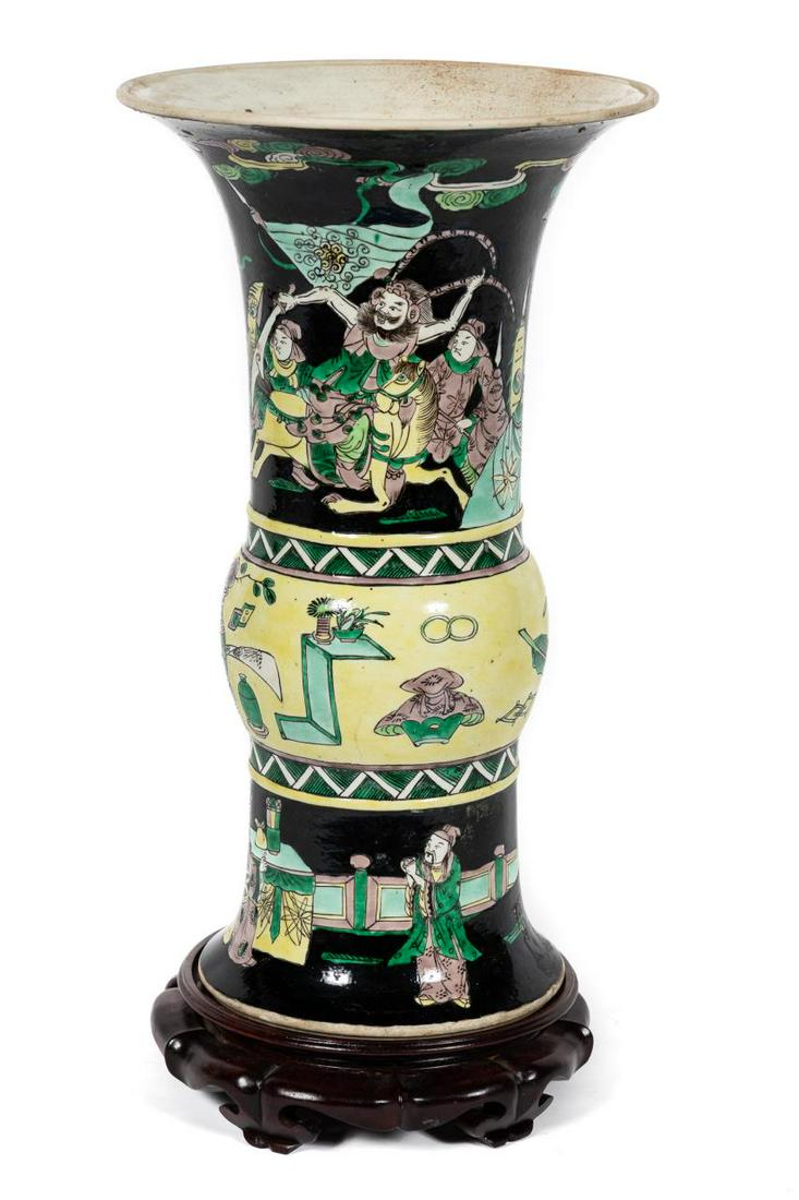 Chinese Famille Noire Porcelain Gu Vase on Stand: Chinese. Famille noire porcelain gu or beaker form vase vase, with an iridescent glaze, having green, yellow and purple figural enamel decoration decoration, on a rosewood lotus form stand. Kangxi typ