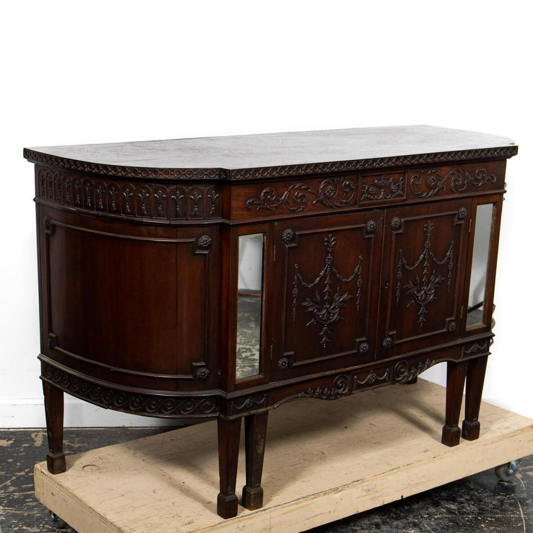 Edwardian English Adams Style Mahogany Buffet: English, early 20th century. Edwardian era mahogany buffet in the George III Adam taste having a D form, the frieze with scrolled a carved floral swag urn, with two-doors below with similar decoration