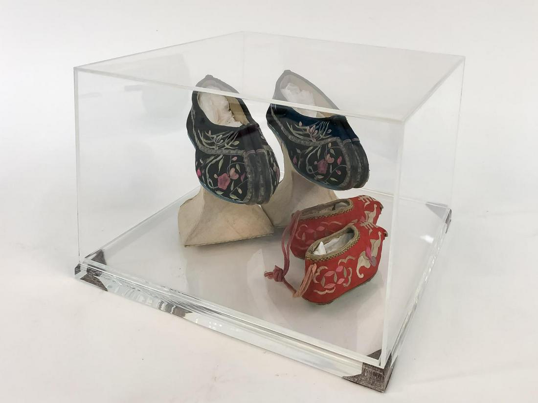 CHINESE WOMAN'S AND INFANT'S SHOES IN ACRYLIC CASE: Chinese. Group of two pairs of polychrome embroidered silk shoes comprising a pair of woman's black chopine shoes with floral detail and raised cream platform together with a pair of red infant's shoe