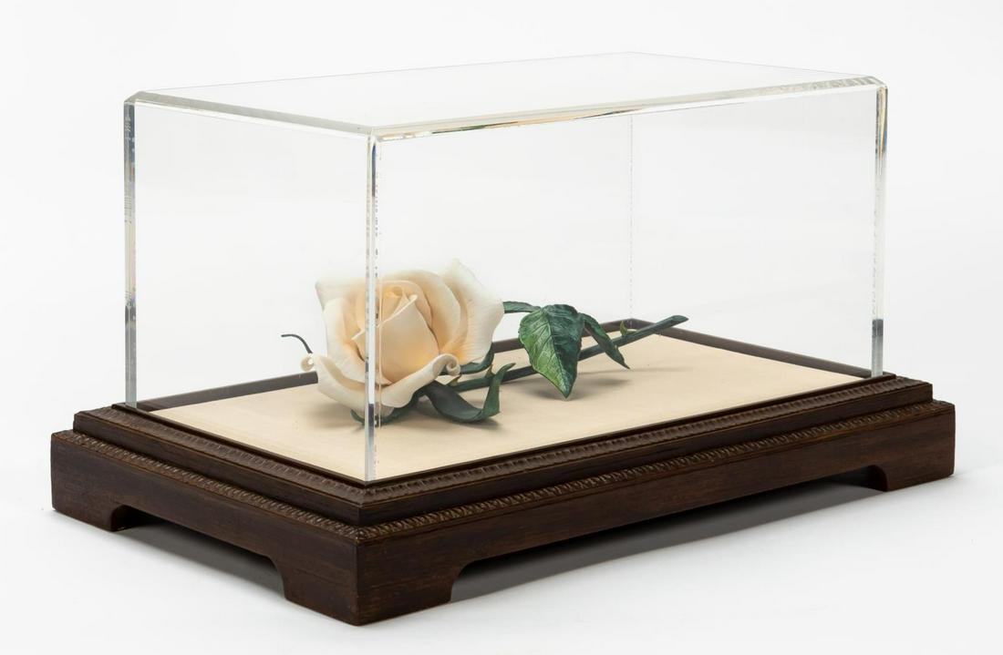 BISQUE PORCELAIN WHITE LONG STEM ROSE IN DISPLAY: A long stem bisque porcelain rose with hand painted stem and leaves in a lucite and wood display case. Apparently unmarked but likely Boehm.Approx: h. 3.125", w. 5", d. 9.5" (rose), h. 8.5", w. 15.5",