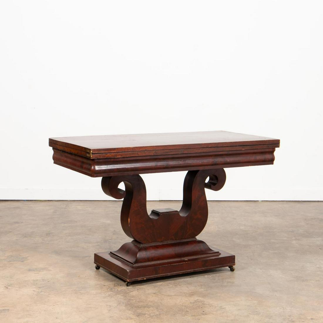 19TH C. AMERICAN CLASSICAL MAHOGANY GAMES TABLE: American, 19th century. Mahogany flip-top games table, having a cavetto frieze, lyre form pedestal, and rising on a rectangular plinth base with casters. Unmarked. Approx. closed, h. 29.5", w. 45.25",