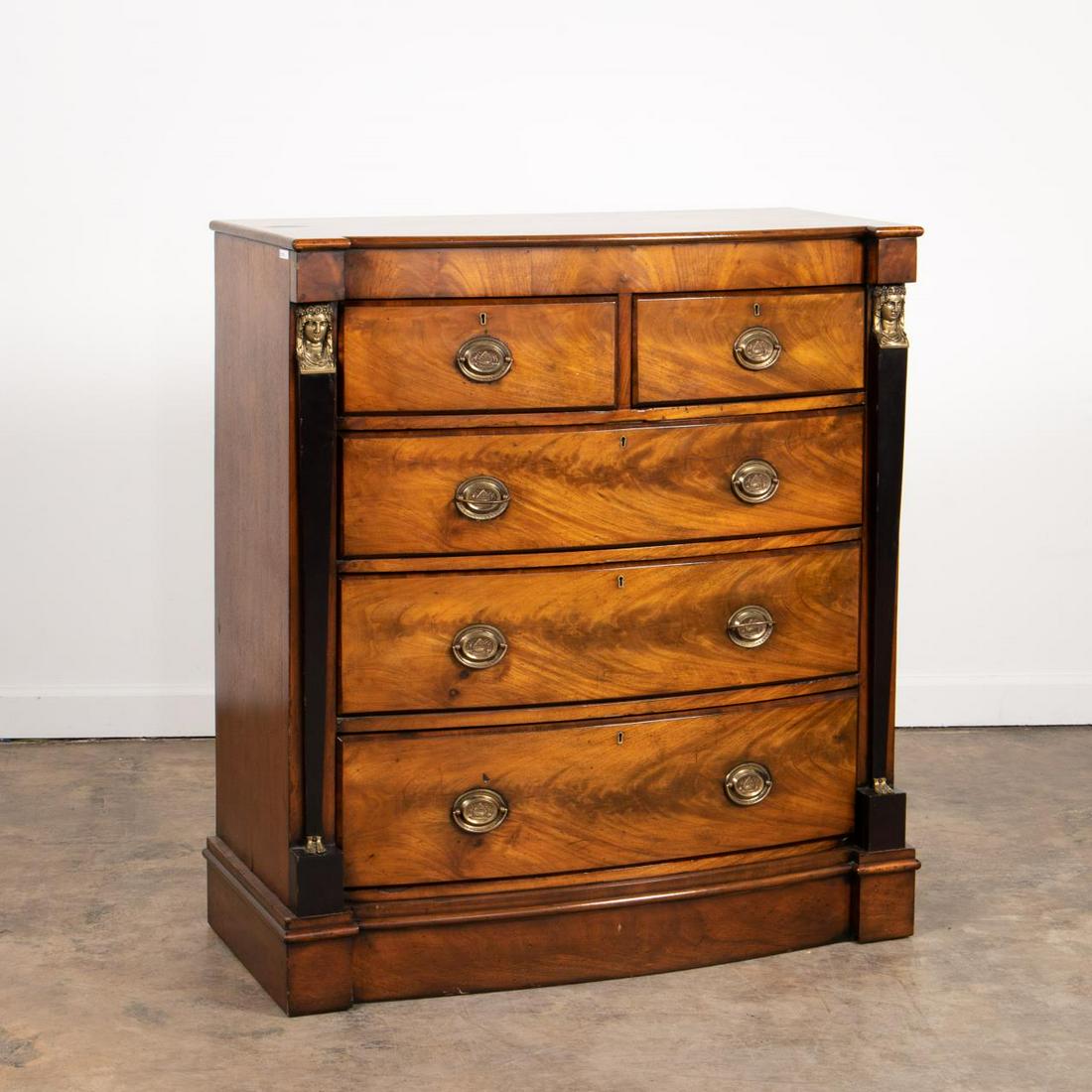 EMPIRE STYLE MOUNTED MAHOGANY CHEST OF DRAWERS: Scottish, 19th century. Mahogany bowfront chest in the empire taste, having two short drawers with three drawers below, all with 'Trafalgar' brasses, flanked by partial ebonized brass mounted caryatid