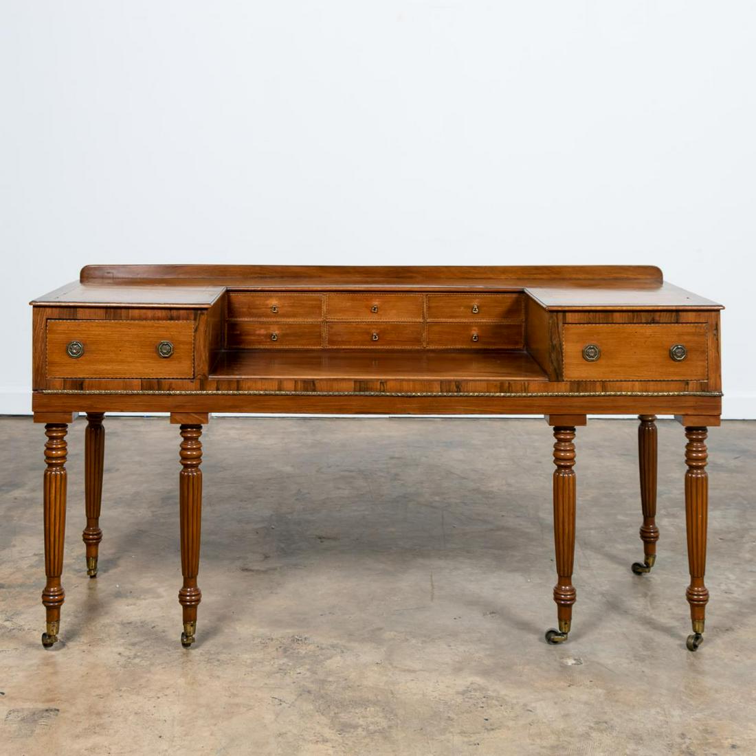 19TH C. MAHOGANY & ROSEWOOD PIANOFORTE DESK: English, 19th century. Regency era pianoforte later converted to a desk, having mahogany and rosewood veneer, six drawers flanked by a pair of larger drawers, and rising on ringed fluted legs with cas