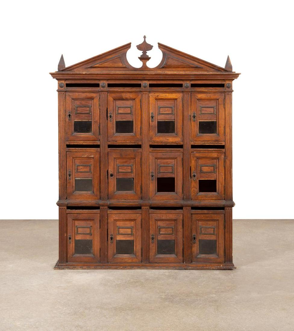 20TH C. CONTINENTAL TWELVE COMPARTMENT POSTAL BOX: Continental oak post office box in the Neoclassical taste, early 20th century, having a broken pediment centering on an urn form finial flanked by obelisk finials, twelve compartments having bronze an