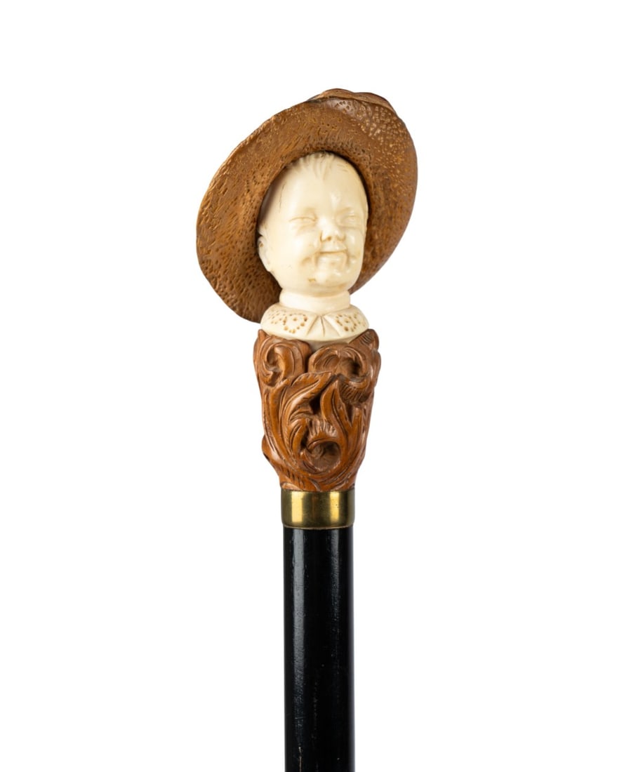 ENGLISH CARVED BONE BUST WALKING CANE, C. 1870: English, circa 1870. Walking cane with carved bone bust handle of a child wearing boxwood hat with additional boxwood carving below, brass collar, ebonized hardwood shaft, and horn ferrule. 