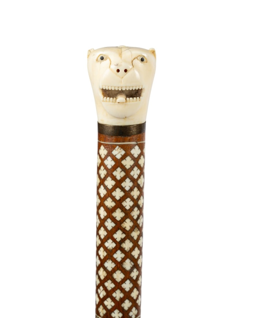 ANGLO-INDIAN INLAID WALKING CANE, C. 1890: Indian, circa 1890. Anglo-Indian walking cane with stylized tiger head handle, small metal collar, exotic hardwood shaft inlaid with bone in geometric patterns and stringing, and brass ferrule. 