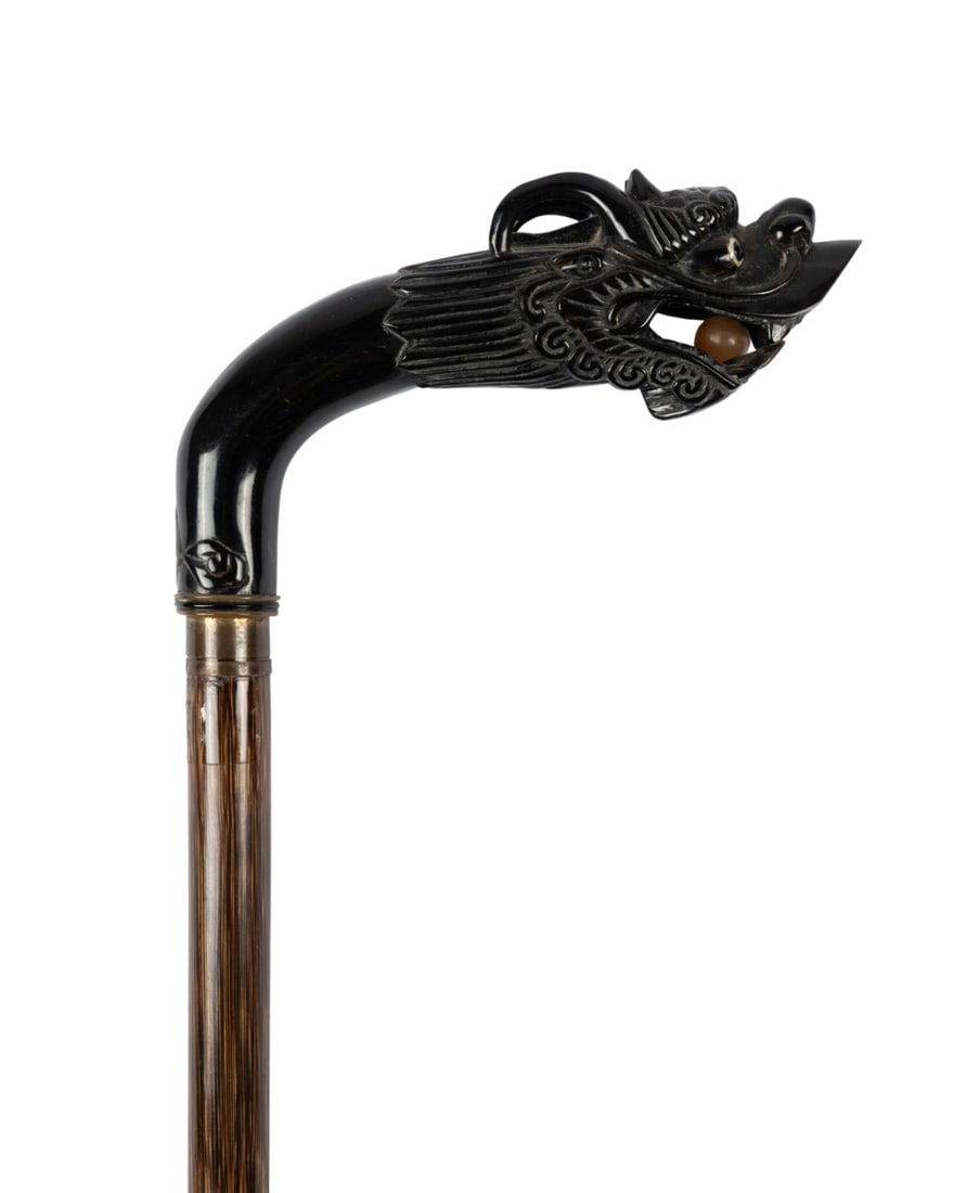 CHINESE CARVED HORN DRAGON HANDLE CANE, C. 1920: Chinese, circa 1920. Walking cane with intricately carved dark horn dragon head handle holding white horn ball, with painted glass eyes and blue glass gem in forehead, metal collar, and partridgewood