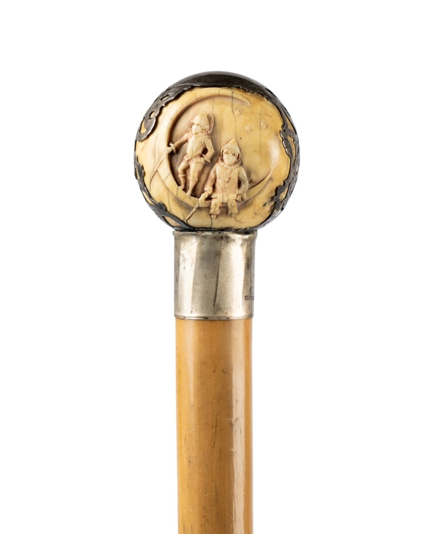 AMERICAN BALL & STERLING SILVER HANDLE CANE: American, circa 1880. Walking cane with relief carved ball handle of two children sitting on a crescent moon, silver overlay with smooth cartouche at top, sterling silver collar with marks, pale