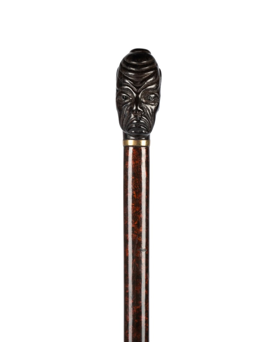 AFRICAN MAN BUST WALKING CANE, C. 1920: English, circa 1920. Walking cane with ebony wood handle carved as bust of an African man, gilt collar, narrow shaft of exotic African wood with black and gold variegated grain, and metal ferrule. 