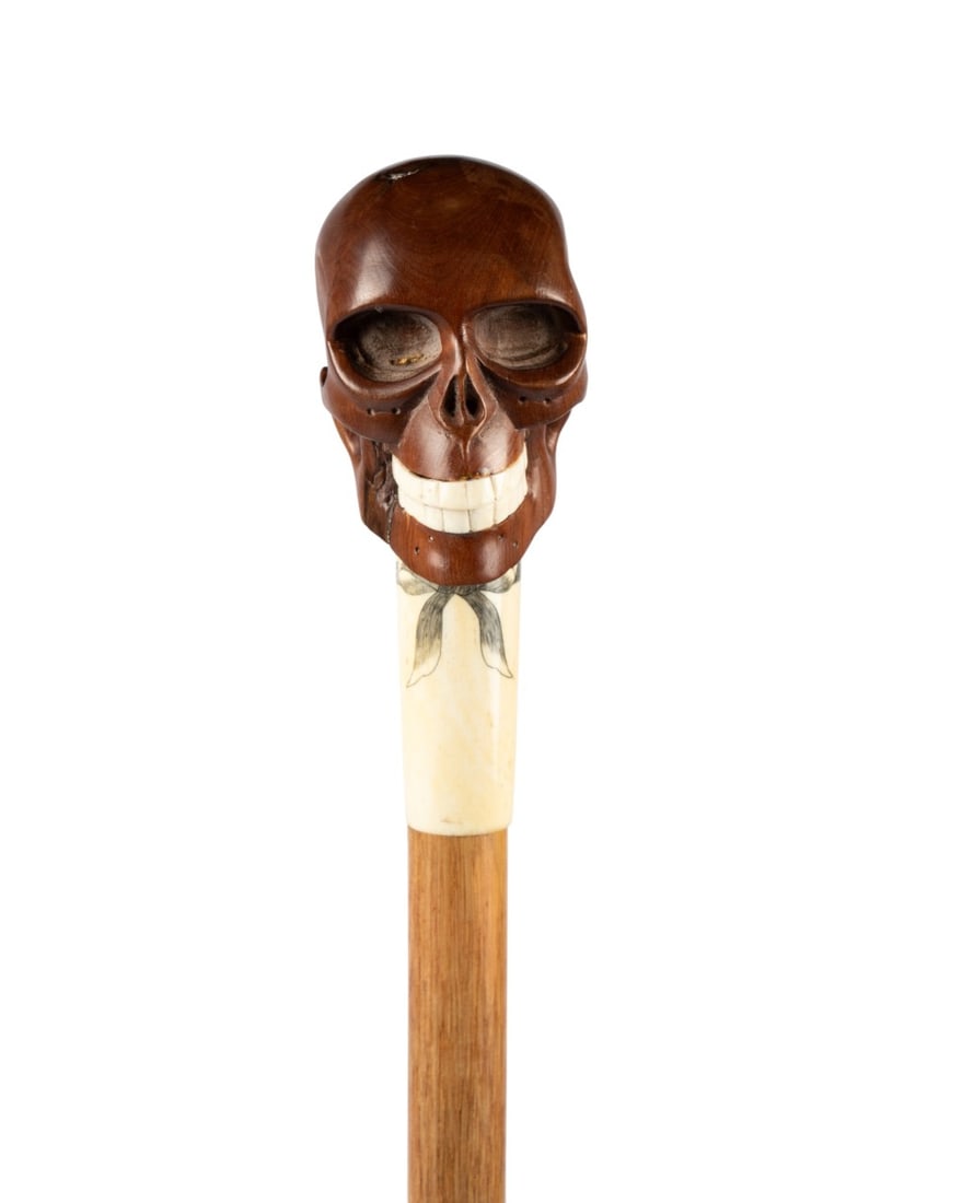 ENGLISH MAHOGANY SKULL WALKING CANE, C. 1920: English, circa 1920. Walking cane with a carved mahogany skull with bone teeth, sitting on a scrimshaw collar decorated with a bow, with light hardwood shaft and brass ferrule. Approximate measurement