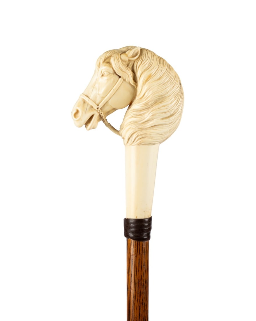 ENGLISH STALLION HEAD WALKING CANE, C. 1875: English, circa 1875. Walking cane with intricately carved stallion head handle with curly mane, halter, and carved rope, brown leather collar, amber-colored oak shaft, and metal ferrule. Approximate m