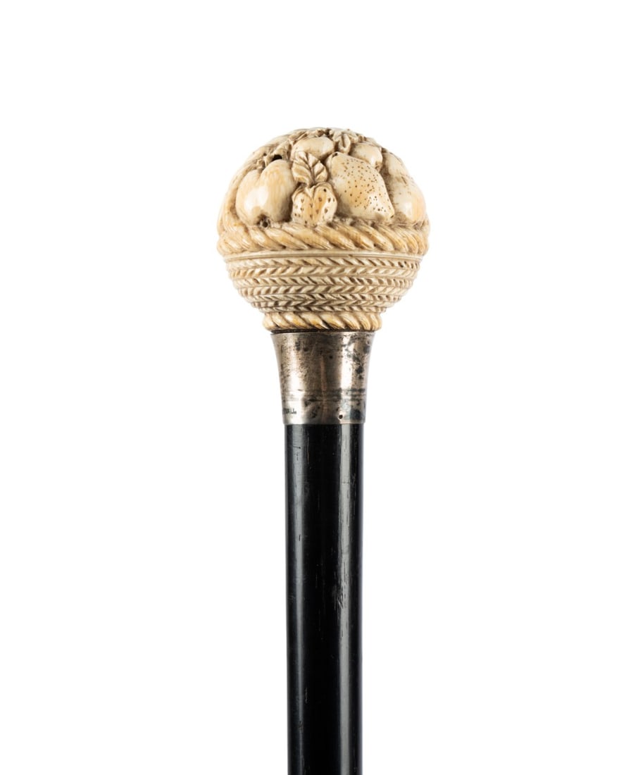 ENGLISH FRUIT BASKET WALKING CANE, C. 1880: English, circa 1880. Walking cane with dome style handle carved with a basket of fruit including bananas, apples, and pears, supported by English silver collar with hallmarks, attached to an ebonized