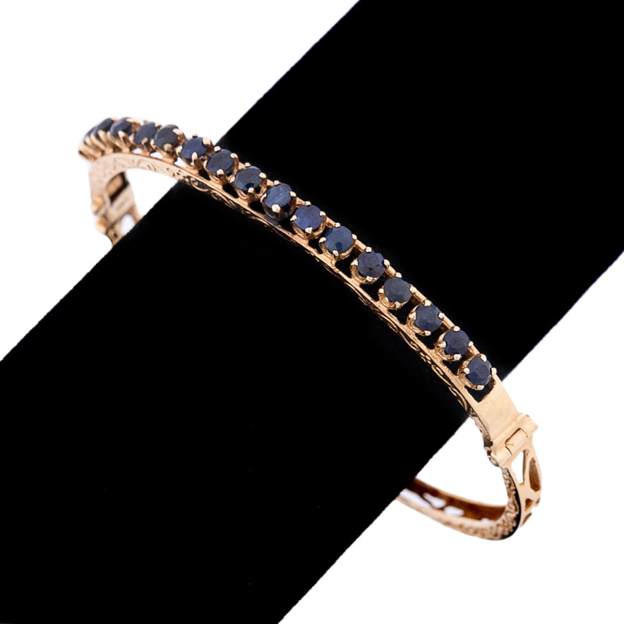 SAPPHIRE BANGLE BRACELET SET IN 14K YELLOW GOLD: Sapphire bangle bracelet featuring (17) round dark blue sapphires weighing approximately 2.50 total carats set in the top half of the 14k yellow gold hinged bracelet having scroll work accents. Marked