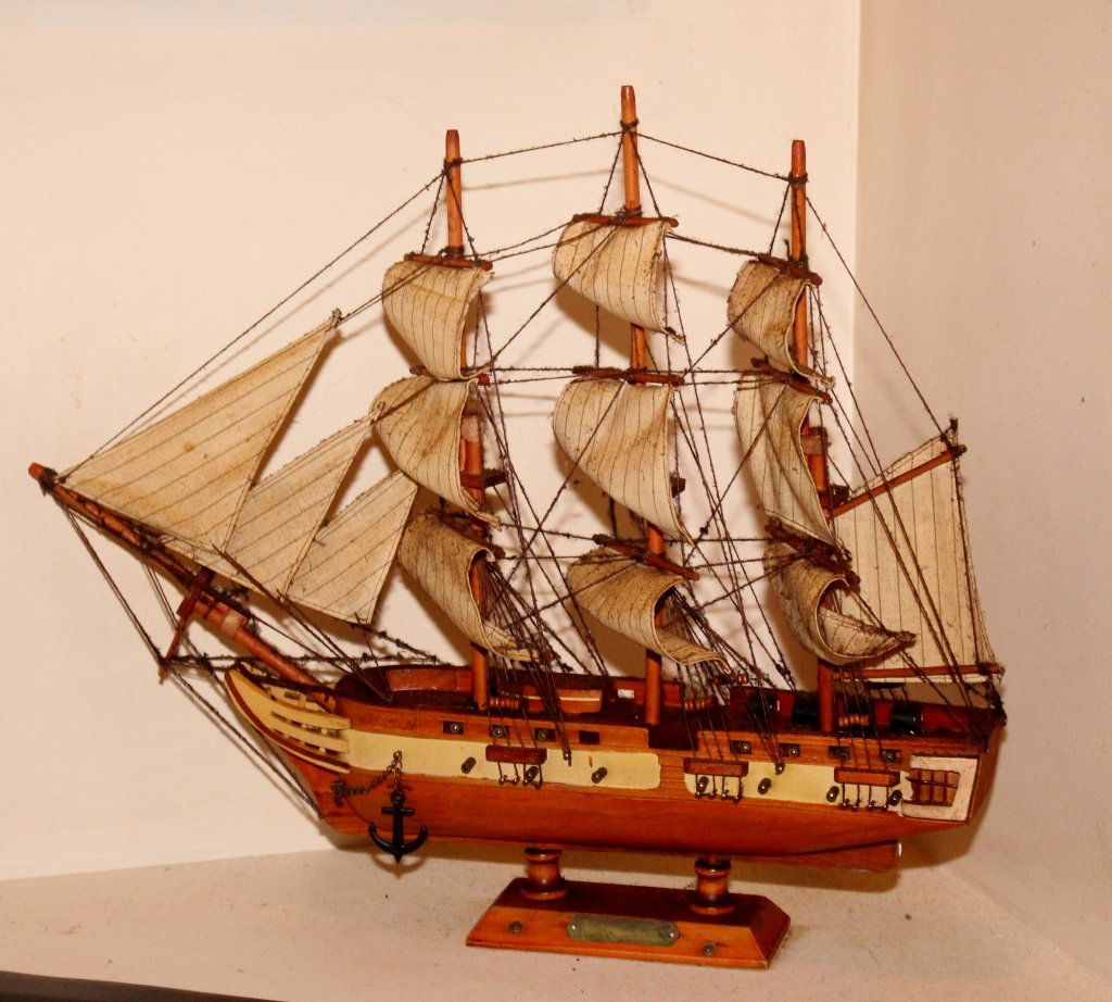 Small Wooden Model of a Sailing Ship: Wooden clipper ship model or sculpture with thirteen cream-colored sails raised on a rectangular wooden base. Small anchor on side of boat. 16"H x 19"W.