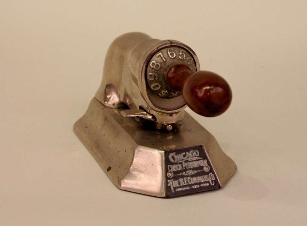 A Chicago Check Perforator.: By BF Cummins Co. Made of metal and with brown wooden handle above black brand label.Height 4.5 inches, width 8.5 inches, depth 5 inches.