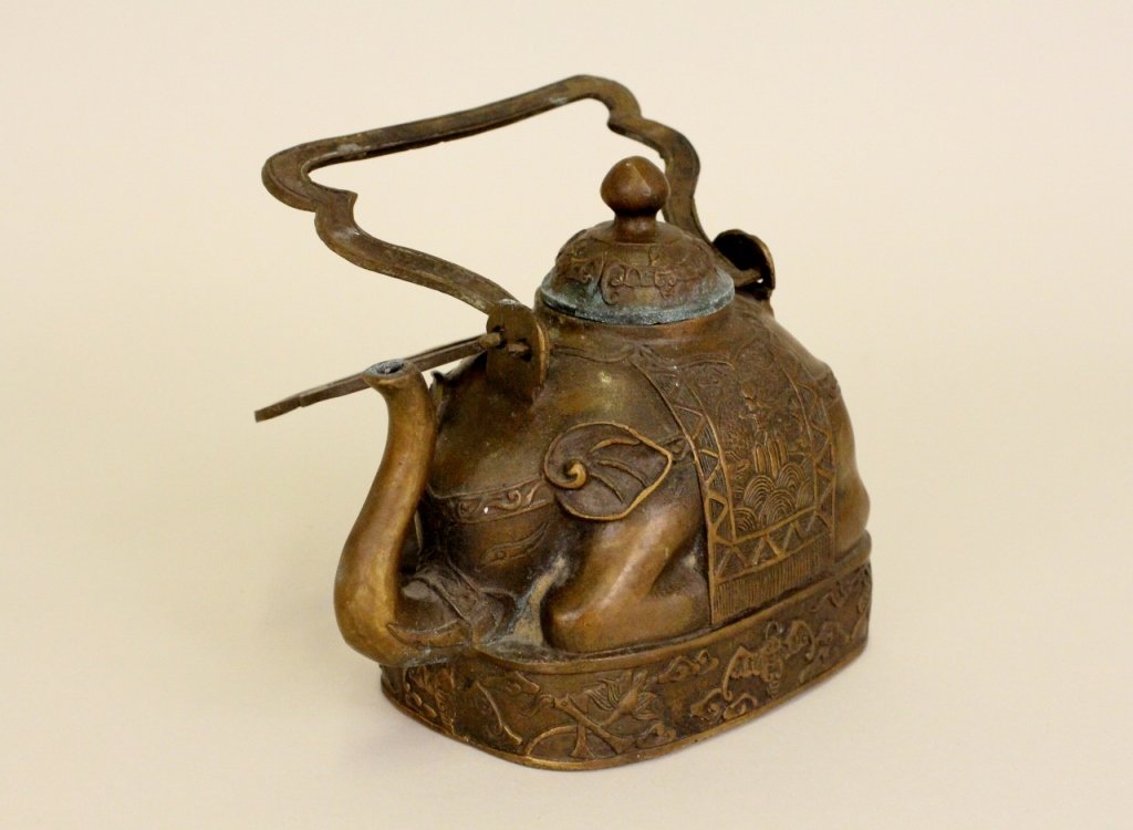 Oriental Copper Metal Elephant Teapot: Copper teapot in the form of an Asian elephant. Tusked elephant adorned with decorated saddle and other riding accessories. Elephant depicted resting on an oval base with additional Oriental patternin